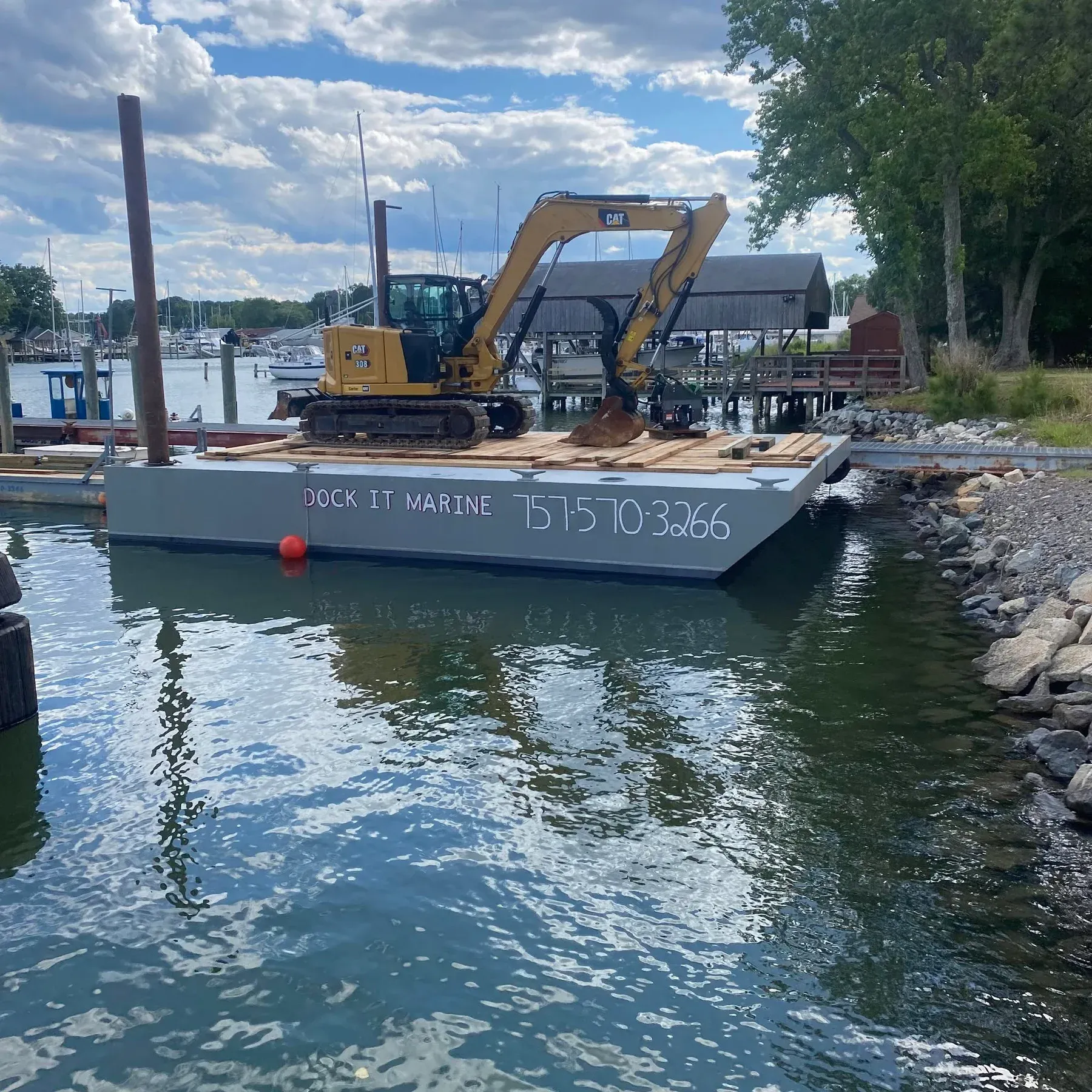 The Dock It Marine barge with DOCK IT MARINE 757-570-3266 painted on the hull and a CAT excavator on deck